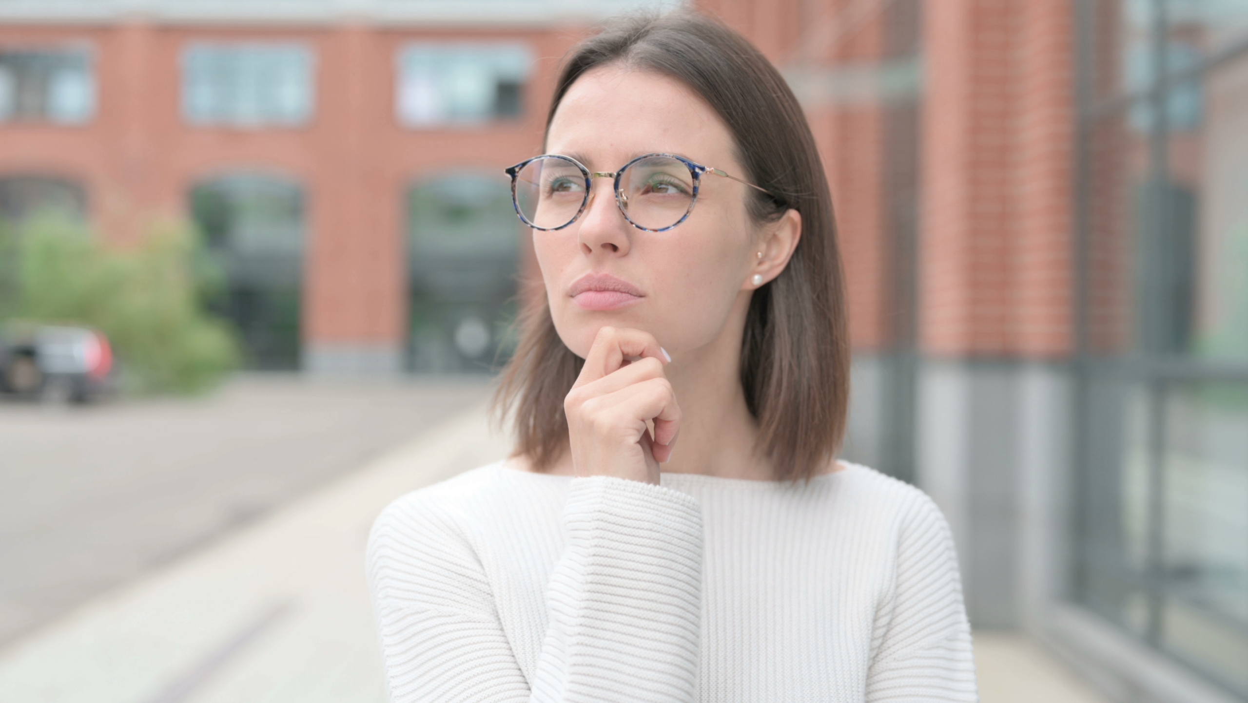 Screenshot of stock footage item. Close up of a white woman with her hand on her chin looking pensively away from the camera. She is wearing glasses and a white jumper.