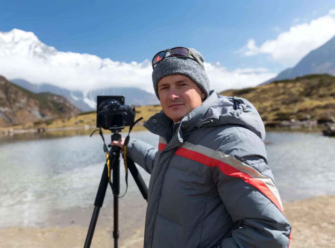 Image of Denys in front of a mountain and lake scene holding a camera on a tripod.
