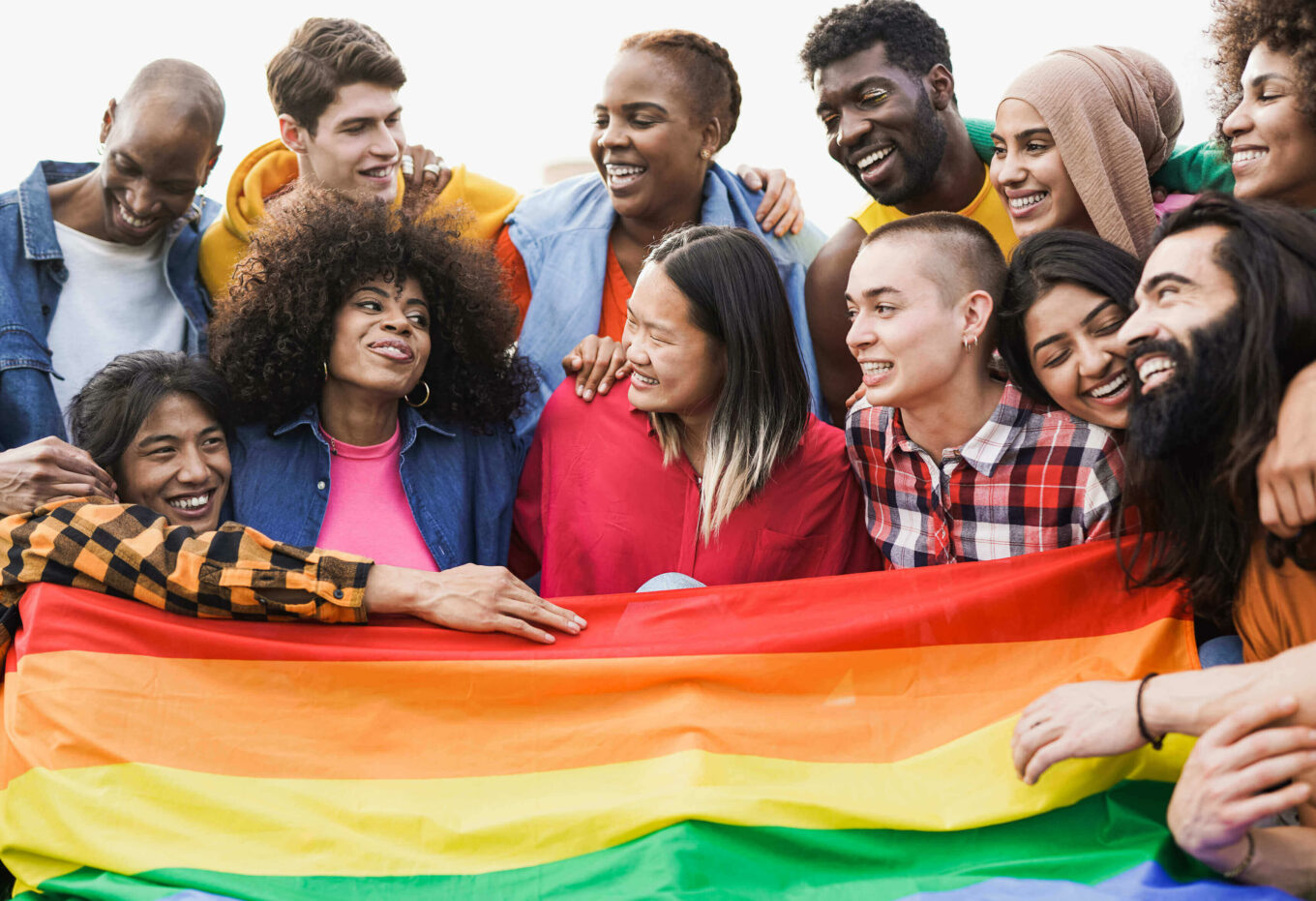 Group of people holding their arms around each other's shoulders while holding a rainbow flag.